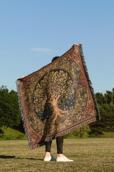 a person standing in a field holding up a large tapestry with a tree on it