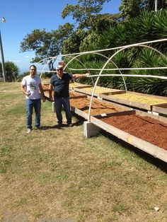 two men standing next to each other in front of a row of wooden troughs filled with dirt