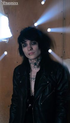 a woman with black hair and piercings standing in front of a wooden wall wearing a leather jacket