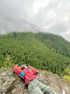 a man laying on top of a rock next to a lush green forest covered hillside