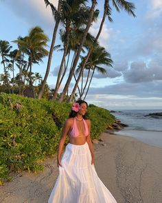 a woman in a white dress standing on the beach with palm trees and water behind her