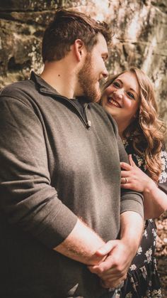 a man and woman standing next to each other in front of a rock wall with their arms around each other