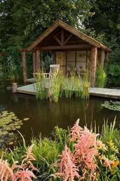 a small wooden structure sitting in the middle of a pond surrounded by flowers and greenery