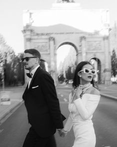 a man and woman are walking down the street in front of an arch on a sunny day