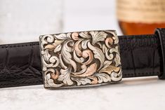 This buckle is made on a hand-wrought sterling silver base with floral patterning composed of 14k rose gold, 14k yellow gold, sterling silver, and silver/copper mokume gane. Mokume gane, Japanese for “wood grain metal," is an alloying technique that fuses several layers of precious metals to produce beautiful multi-color blending and rippling patterns. This is a wonderful example of mokume gane—oftentimes the patterns are almost too small to see with the naked eye, but expert technique and cleve Cowboy Spurs, Leather Belt Buckle, Mokume Gane, Leather Carving, Buckle Bracelet, Metal Engraving, Color Blending, Men's Style, Belt Buckle