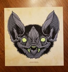 a drawing of a bat with glowing eyes and fangs on it's face, sitting on a wooden surface