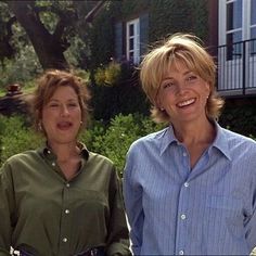 two women standing next to each other in front of a house with trees and bushes