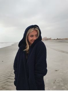 a woman standing on top of a sandy beach next to the ocean wearing a blue coat