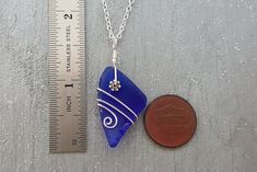 a penny is next to a blue sea glass pendant on a silver chain with a measuring ruler behind it