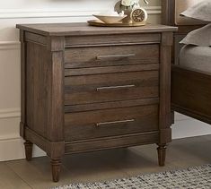 a night stand with three drawers and a clock on the top, next to a bed