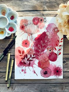 watercolor flowers on paper next to paints and paintbrushes sitting on a wooden table