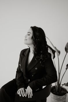 a woman in a suit sitting on a chair next to a potted plant and looking off into the distance