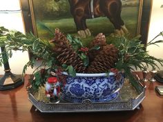 a blue and white bowl filled with pine cones on top of a wooden table next to a painting