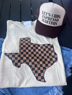 Experience the rugged spirit of Texas with our Checkered Texas Tee! Made with a classic checkered design, this tee is perfect for any adventure. Whether you're exploring the great outdoors or simply showing off your Texan pride, our tee will keep you stylish and comfortable. Get yours today! professionally printed tee brand: Comfort Colors color: Ivory comfy, unisex fit ships within 10-14 business days. Casual Gingham Crew Neck Top, Casual Brown Tops For Outdoor Activities, Casual Plaid T-shirt With Graphic Print, Casual Plaid T-shirt For Fall, Graphic Print T-shirt For Fall Camping, Checkered Design, Football Tees, First Halloween, Top Graphic Tees