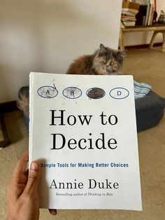 a person holding up a book about how to decide with a cat in the background