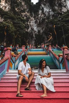 a man and woman sitting on the steps in front of stairs with mountains behind them