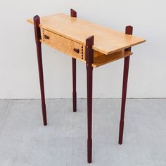 a small wooden desk with two legs and a drawer on the top, in front of a white wall