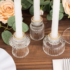 two clear candlesticks sitting on top of a wooden table next to white plates and silverware