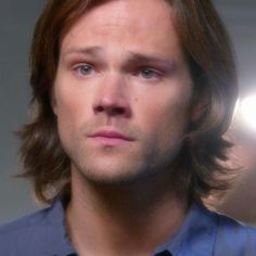 a man with long hair and blue shirt looking off to the side in front of a spotlight