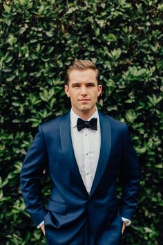 a man in a blue suit and bow tie standing next to a green hedge wall