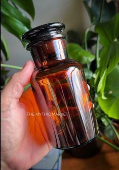 a hand holding an orange glass bottle in front of a potted plant