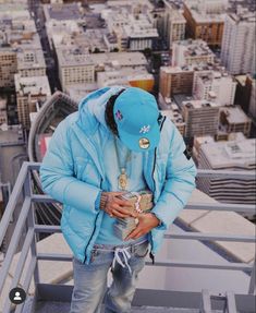 a man standing on top of a tall building wearing a blue jacket and hat with his hands in his pockets