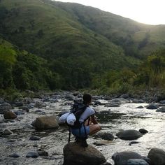 a person sitting on rocks in the middle of a river with mountains in the background