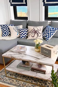 a living room with a couch, coffee table and books on it in front of two windows