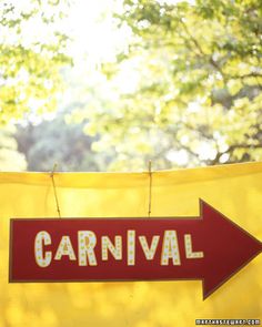 a carnival sign hanging from the side of a yellow tent with trees in the background