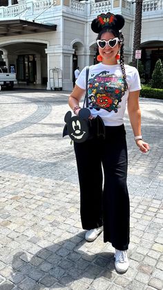 Julia Vaness wearing black linen pants with a white graphic tshirt from Disneyland 2023 Halloween collection, with loungefly mickey mouse bat bag, pumpkin plush earrings, white heart shaped sunglasses and black fuzzy halloween mickey ears