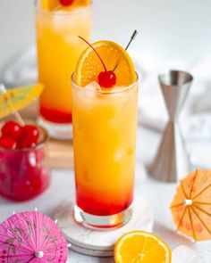 two glasses filled with orange juice and garnished with cherries next to umbrellas