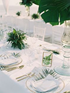 the table is set with white linens and gold place settings, greenery in glass vase