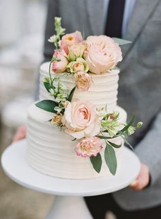 a white wedding cake with pink flowers on top and greenery around the edges is being held by a man in a gray suit