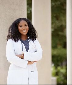 a woman wearing a white lab coat and standing with her arms crossed in front of columns