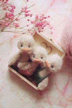 three small stuffed animals in a box on a pink marble surface with flowers and yarn