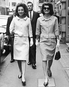 two women walking down the street with men in suits behind them and one woman wearing sunglasses