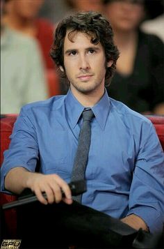 a man wearing a blue shirt and tie sitting in front of a red chair with his hands on the steering wheel