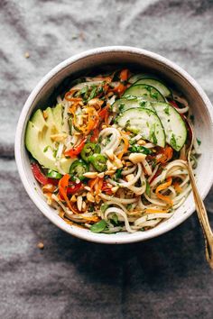 a white bowl filled with noodles, cucumbers and other veggies next to a spoon