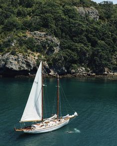 a sailboat in the water near some rocks and green trees on top of a hill