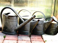 three metal watering cans sitting on top of a brick floor