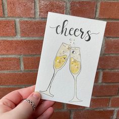 a hand holding up a card with cheers written on it and two champagne flutes in front of a brick wall