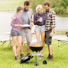 three people cooking food on a grill in the grass near a lake and picnic tables