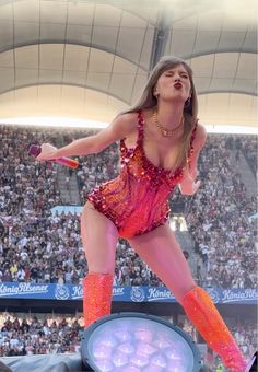 a woman in a pink and orange outfit on stage with an audience watching from the bleachers