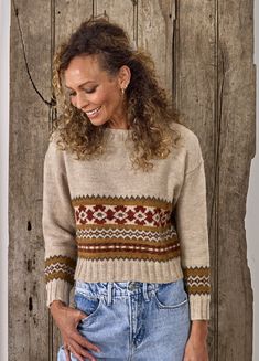 a woman standing in front of a wooden wall wearing a sweater and jeans with her hands on her hips