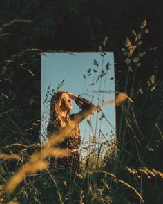 a woman standing in tall grass with her hand on her head looking at the sky