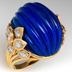 This exquisite cocktail ring is centered with one (1) oval shaped natural lapis lazuli with a carved fluted design. The shoulders of the ring are each accented with six (6) round brilliant cut diamonds bead set into petal shaped setting. The ring measures 23.6mm at the top, rises 15.8mm above the finger, tapering to 2.6mm wide and 1.2mm thick at the base of the shank. It is currently a size 7.25. Luxury Lapis Lazuli Engagement Rings, Luxury Formal Blue Turquoise Ring, Luxury Classic Domed Sapphire Ring, Luxury Gold Sapphire Fusion Ring, Elegant Lapis Lazuli Ring With Polished Finish, Elegant Lapis Lazuli Rings For Formal Occasions, Elegant Formal Lapis Lazuli Rings, Carved Stone Jewelry, 2024 Jewelry