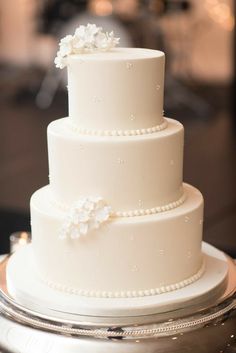 a three tiered white wedding cake with pearls and flowers on the top, sitting on a silver platter