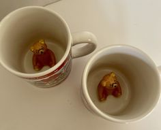 two coffee mugs with small bears in them on a white counter top, one is filled with water