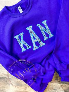 a purple sweatshirt with the letters k and m on it, sitting on a wooden floor