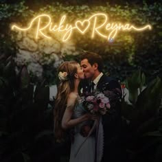 a man and woman kissing in front of a neon sign that reads riley & ryan